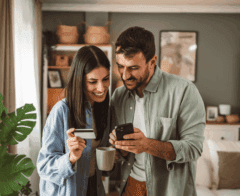 A smiling couple stands together in their living room, looking at a smartphone. The woman holds a credit card and a coffee mug, while the man points at the phone screen. They appear engaged and happy, suggesting they are making an online donation or purchase.