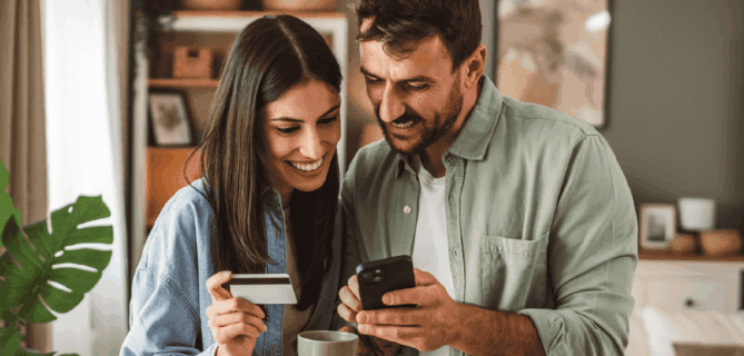A smiling couple stands together in their living room, looking at a smartphone. The woman holds a credit card and a coffee mug, while the man points at the phone screen. They appear engaged and happy, suggesting they are making an online donation or purchase.