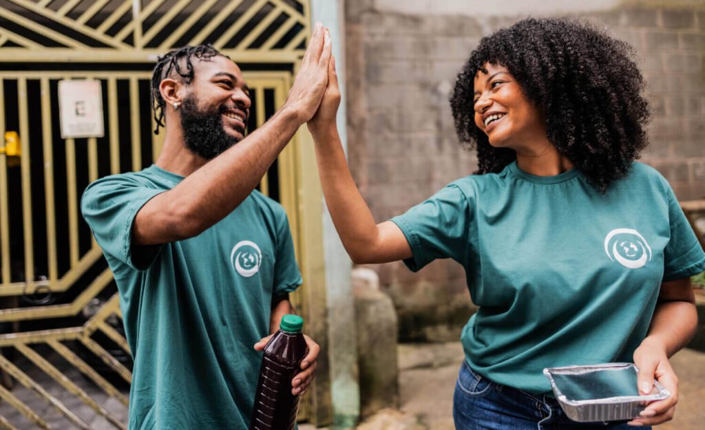 Two volunteers wearing matching green T-shirts smile and give each other a high-five while standing outdoors. One person holds a drink bottle, and the other carries a foil food container. The scene conveys teamwork and positivity during a community service activity.
