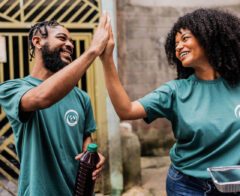 Two volunteers wearing matching green T-shirts smile and give each other a high-five while standing outdoors. One person holds a drink bottle, and the other carries a foil food container. The scene conveys teamwork and positivity during a community service activity.