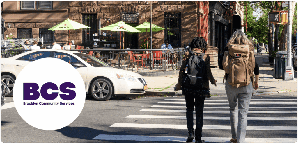 A city street scene in Brooklyn with two people crossing at a crosswalk, backpacks on. A white circle overlays the bottom left with the purple logo: “BCS Brooklyn Community Services.”