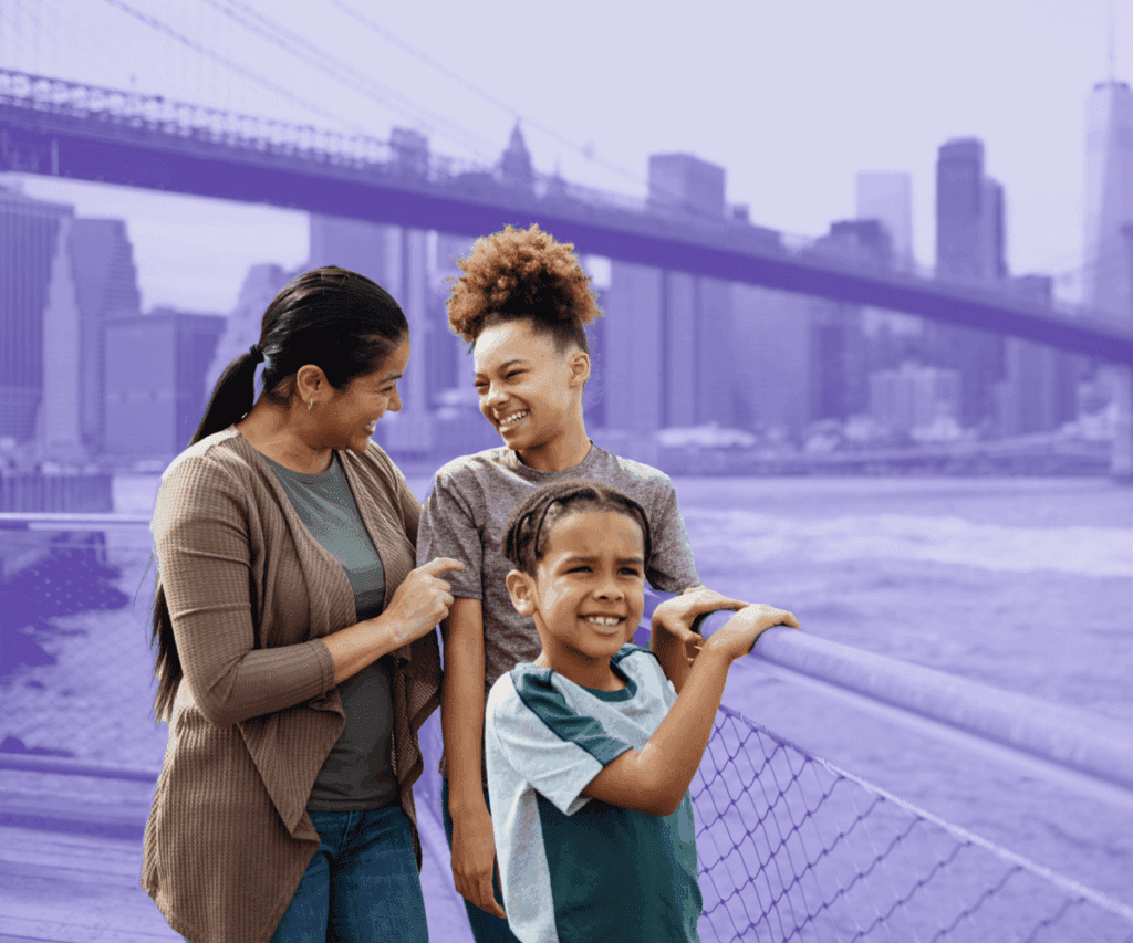 A smiling family of three — a woman, a young girl, and a young boy — stand together outdoors by the waterfront with the Brooklyn Bridge and New York City skyline in the background, tinted with a purple overlay.