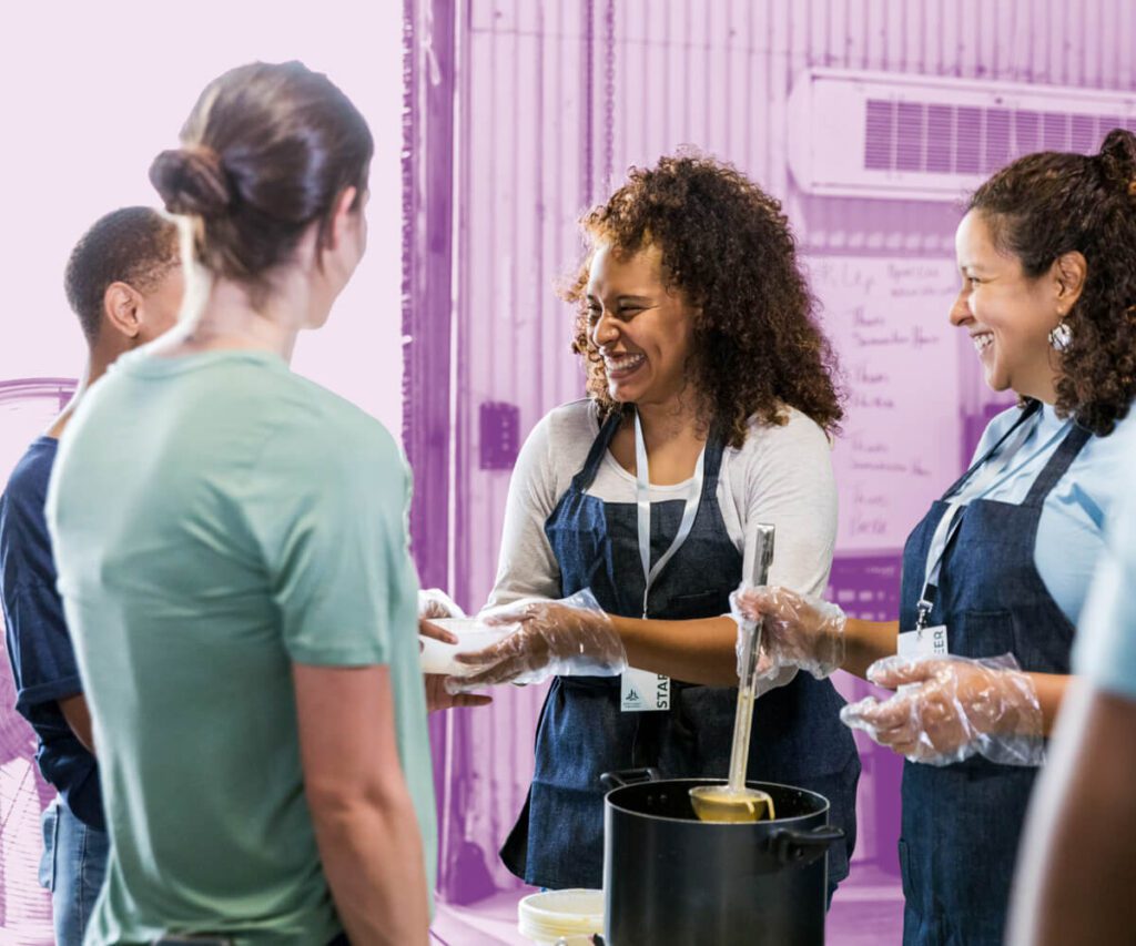 Volunteers at a soup kitchen, wearing aprons and gloves, smiling as they serve food to community members. The background has a purple overlay.