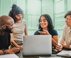 Four colleagues gather around a laptop in a bright office. A woman gestures while explaining something, and the group listens attentively with notebooks and pens in hand.Four colleagues gather around a laptop in a bright office. A woman gestures while explaining something, and the group listens attentively with notebooks and pens in hand.