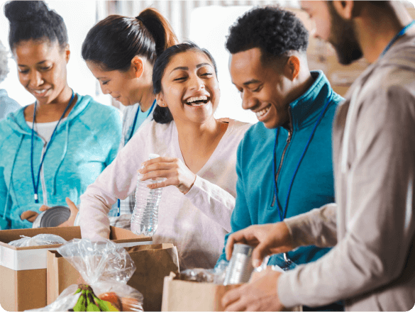 Volunteers at a food drive, smiling and laughing as they pack boxes with bottled water and canned goods. The group appears cheerful and collaborative.