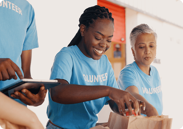 A group of diverse volunteers wearing blue "VOLUNTEER" shirts are smiling and working together. One woman is placing items into a paper bag, while another volunteer stands beside her. In the foreground, another person is holding a tablet. The scene appears to be at a community service or food distribution event.