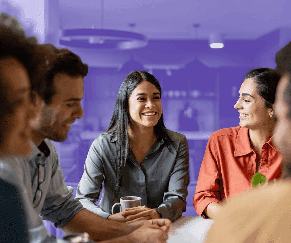 A diverse group of five people are sitting at a table in a brightly lit room, engaged in a cheerful conversation. The woman in the center is smiling broadly. The background is partially covered with a purple overlay, giving emphasis to the people in the foreground.