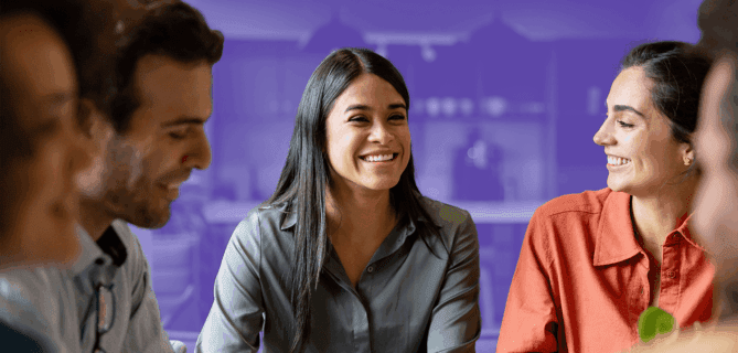 A diverse group of five people are sitting at a table in a brightly lit room, engaged in a cheerful conversation. The woman in the center is smiling broadly. The background is partially covered with a purple overlay, giving emphasis to the people in the foreground.