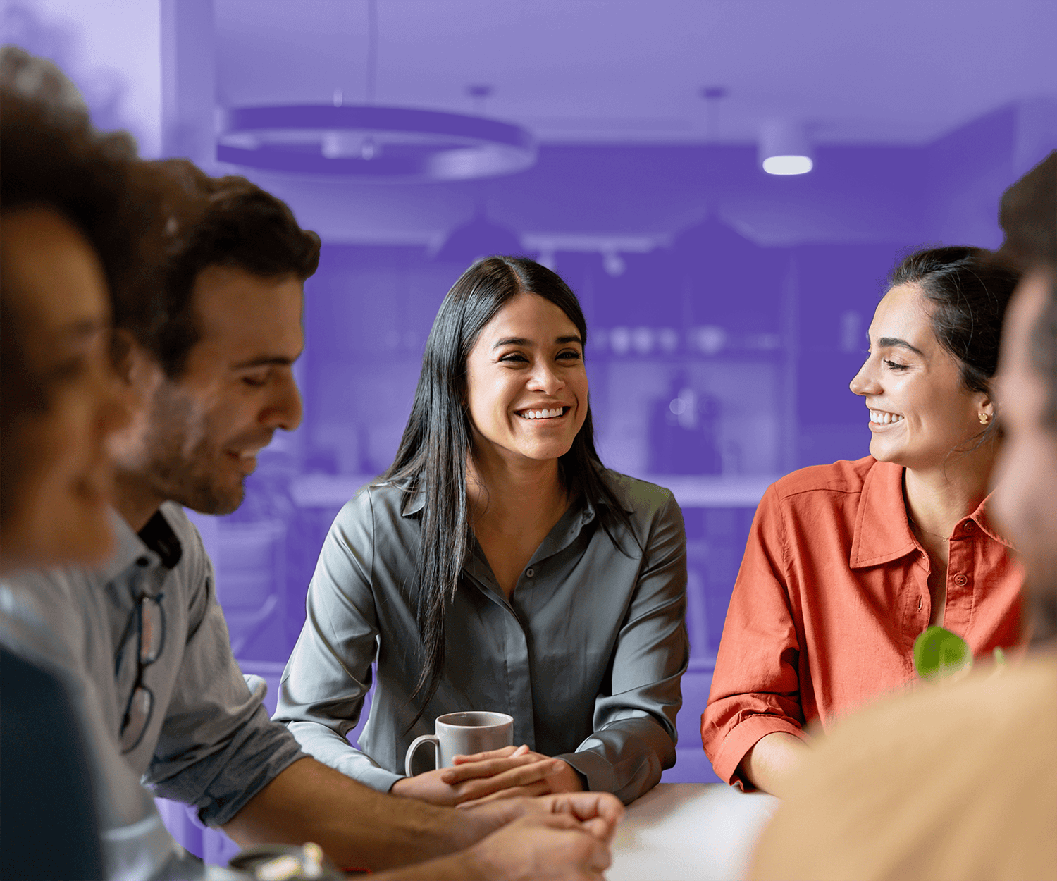 A diverse group of five people are sitting at a table in a brightly lit room, engaged in a cheerful conversation. The woman in the center is smiling broadly. The background is partially covered with a purple overlay, giving emphasis to the people in the foreground.