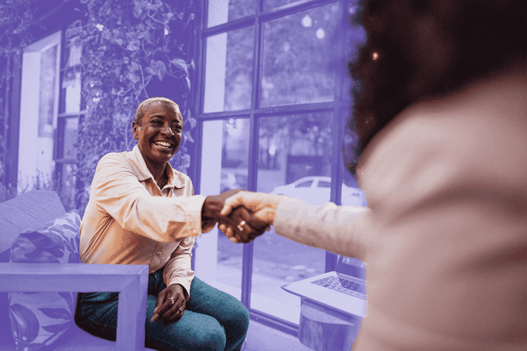 A smiling older woman in a light shirt and teal pants shakes hands with another person across a table in a bright, modern space with large windows and green plants. The background is tinted with a purple overlay, and a laptop sits open on the table beside them.A smiling older woman in a light shirt and teal pants shakes hands with another person across a table in a bright, modern space with large windows and green plants. The background is tinted with a purple overlay, and a laptop sits open on the table beside them.