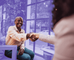 A smiling older woman in a light shirt and teal pants shakes hands with another person across a table in a bright, modern space with large windows and green plants. The background is tinted with a purple overlay, and a laptop sits open on the table beside them.A smiling older woman in a light shirt and teal pants shakes hands with another person across a table in a bright, modern space with large windows and green plants. The background is tinted with a purple overlay, and a laptop sits open on the table beside them.