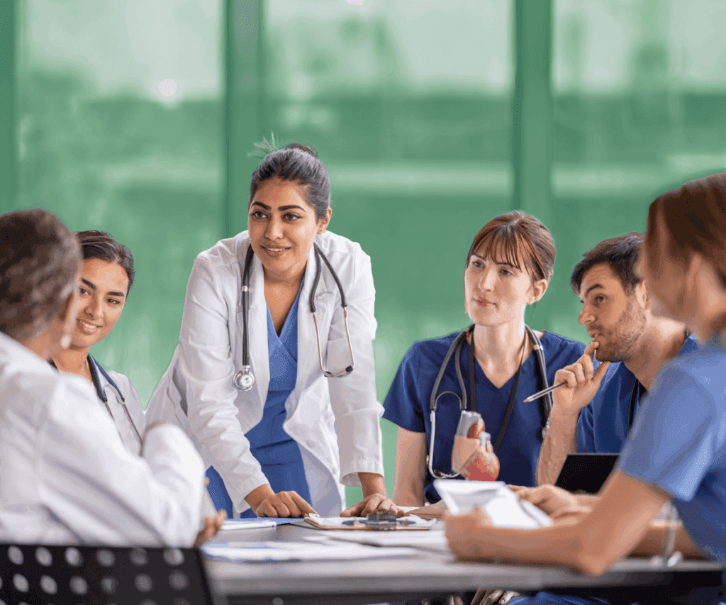 A group of healthcare professionals in scrubs and white coats sit around a table in discussion. A female doctor in the center stands and leans forward, speaking to her colleagues. The background is softly blurred with a green tint.