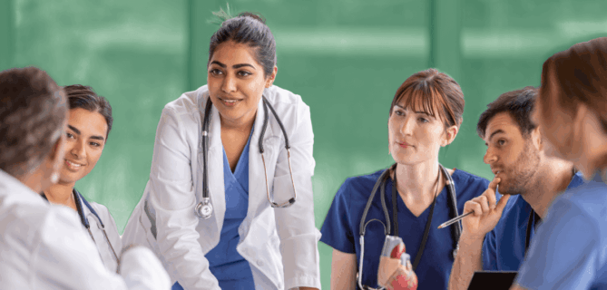 A group of healthcare professionals in scrubs and white coats sit around a table in discussion. A female doctor in the center stands and leans forward, speaking to her colleagues. The background is softly blurred with a green tint.