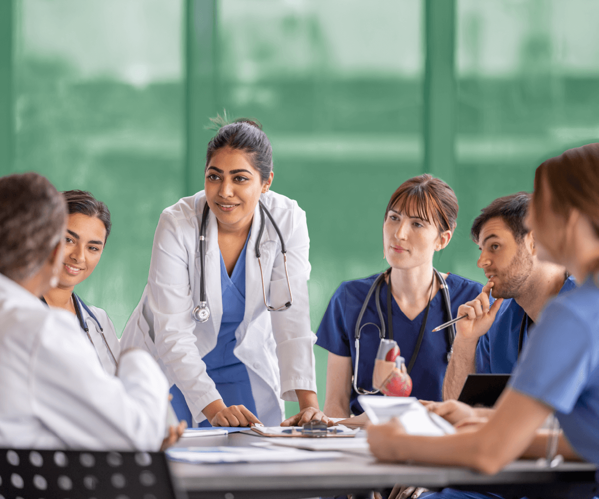 A group of healthcare professionals in scrubs and white coats sit around a table in discussion. A female doctor in the center stands and leans forward, speaking to her colleagues. The background is softly blurred with a green tint.