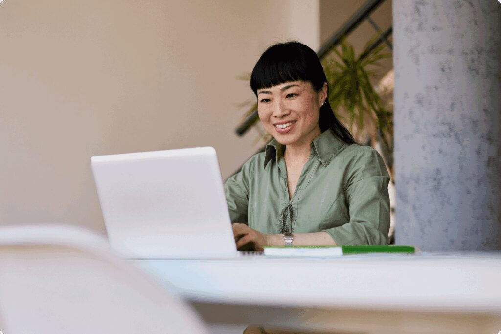 A woman in a green blouse works on a laptop at a desk, smiling as she types. A notebook sits nearby, and the background shows a bright office space with plants.