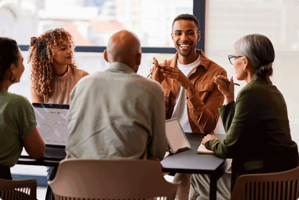 A group of five professionals sit together at a conference table in discussion. One man smiles as he speaks, while others listen and take notes. A laptop with charts is visible, highlighting teamwork and strategy.