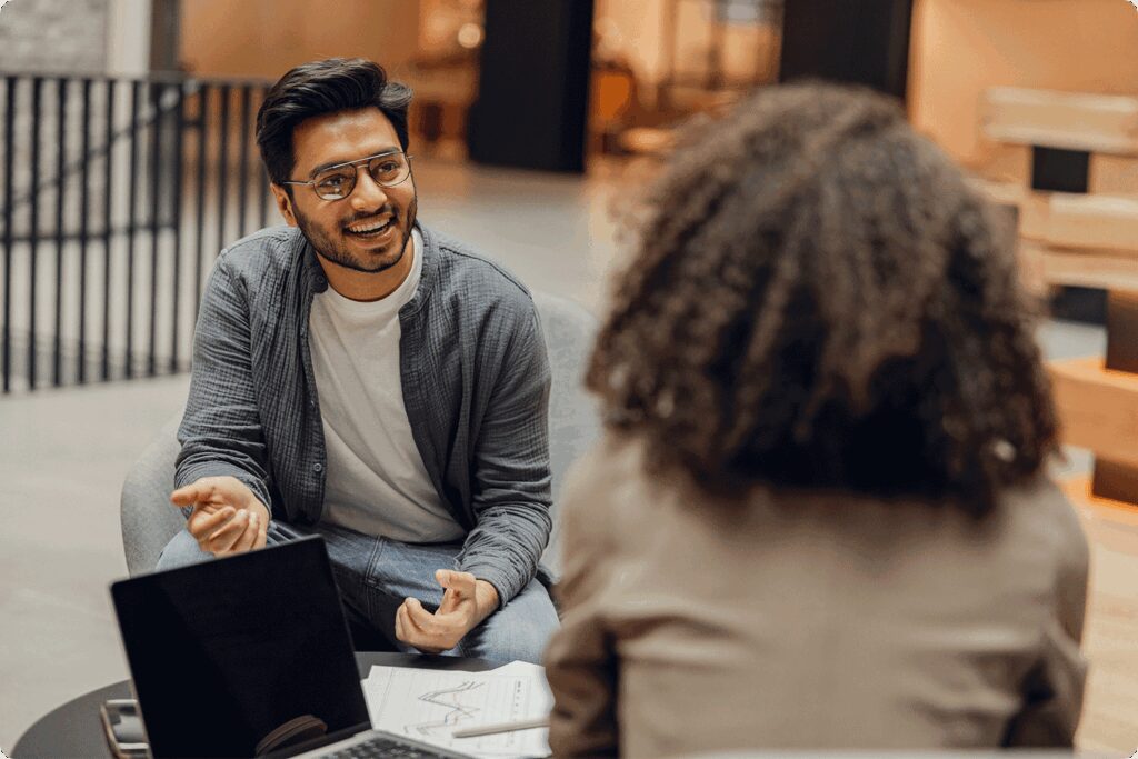 A smiling man wearing glasses sits across from a colleague during a discussion. A laptop with charts and papers rests on the table between them, suggesting a meeting about data or decision-making.