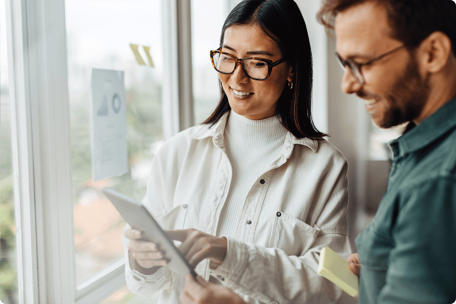Two colleagues stand by a window, smiling while reviewing a tablet together. Papers with charts are taped to the window, suggesting evaluation and data review.