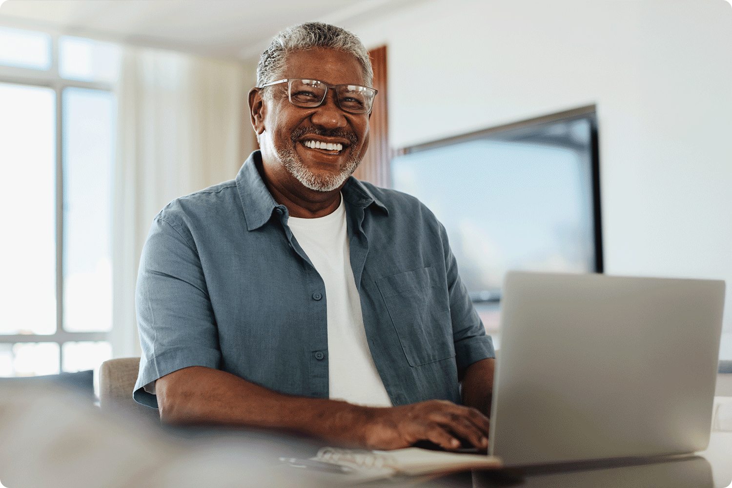 A man in glasses smiles while talking with a colleague across a table. A laptop and printed charts are in front of him, suggesting a discussion about data or workflow improvements.
