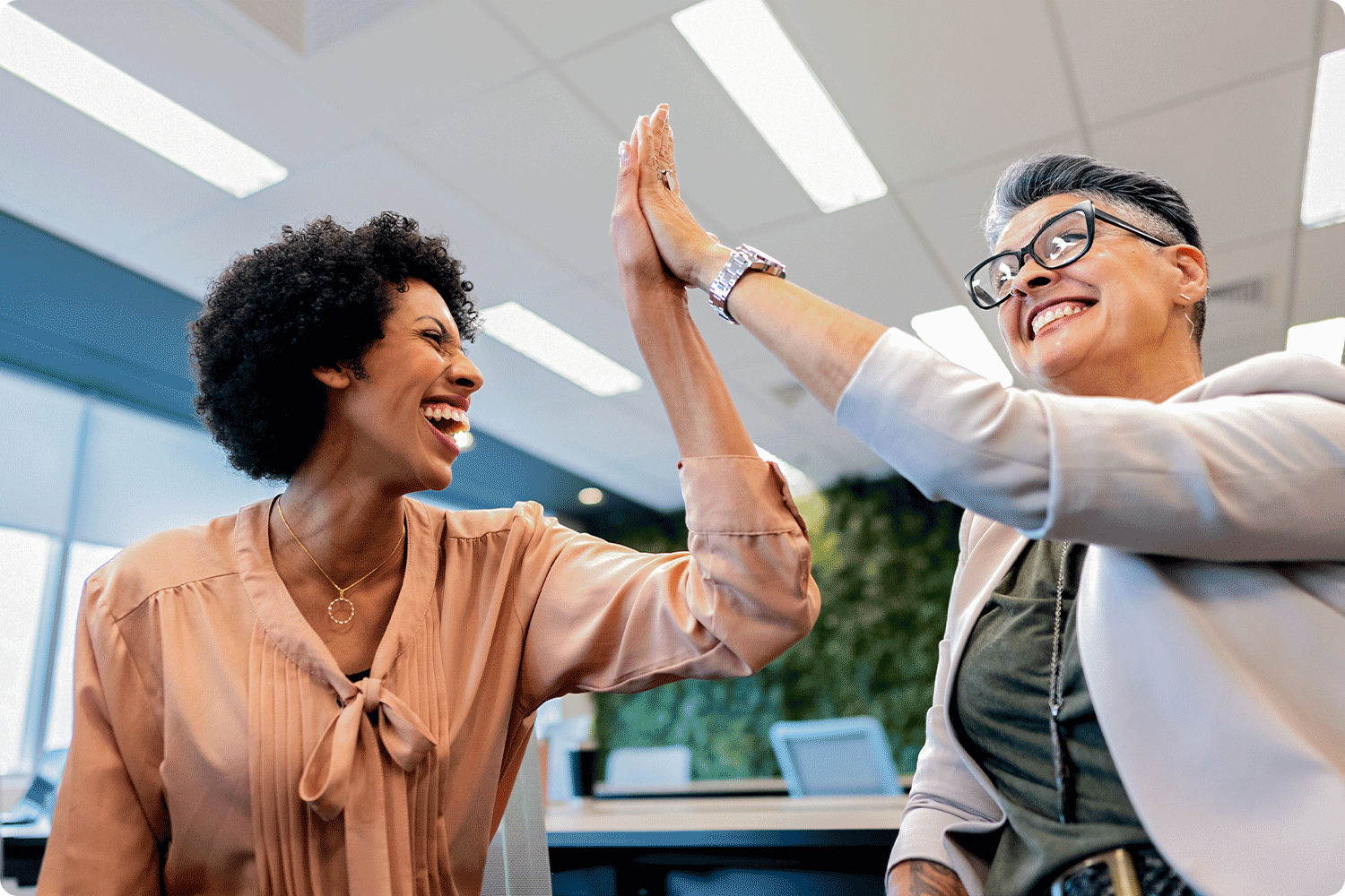 Two women in a professional office setting smile and share a high-five, symbolizing strong partnerships and successful collaboration.