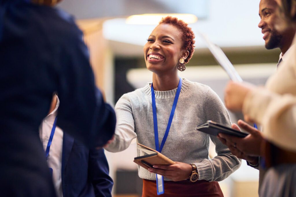 A woman wearing a lanyard smiles as she shakes hands with another person in a professional setting. She holds a notebook, and colleagues around her hold tablets, suggesting a collaborative work environment.