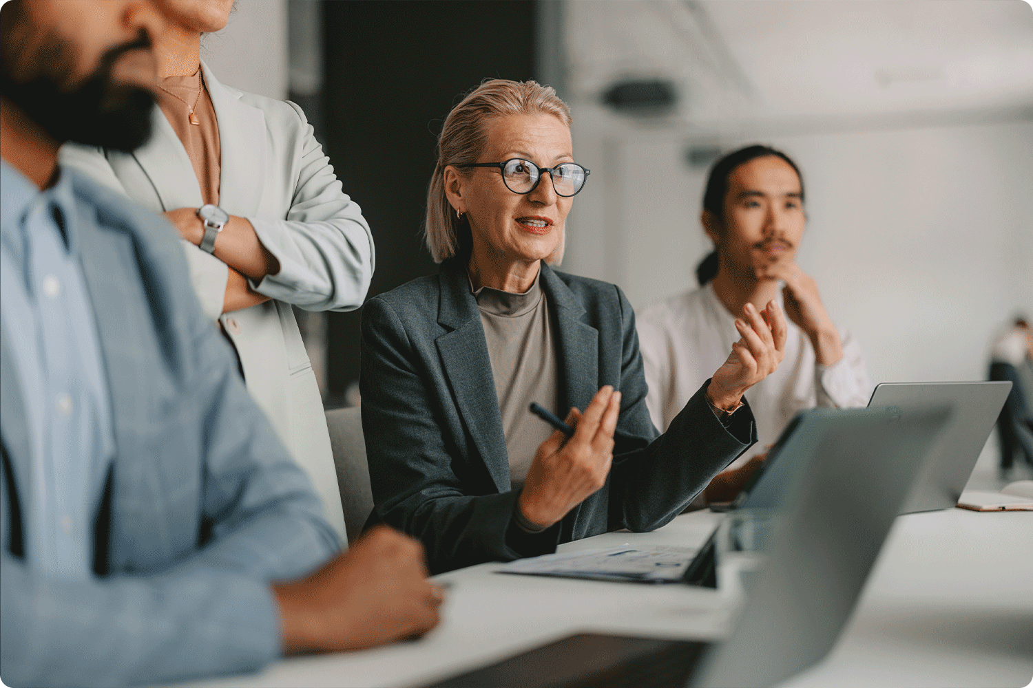 A group of professionals in business attire sit around a conference table. A woman speaks while gesturing with a pen, as others listen attentively, representing accountability and transparent decision-making.