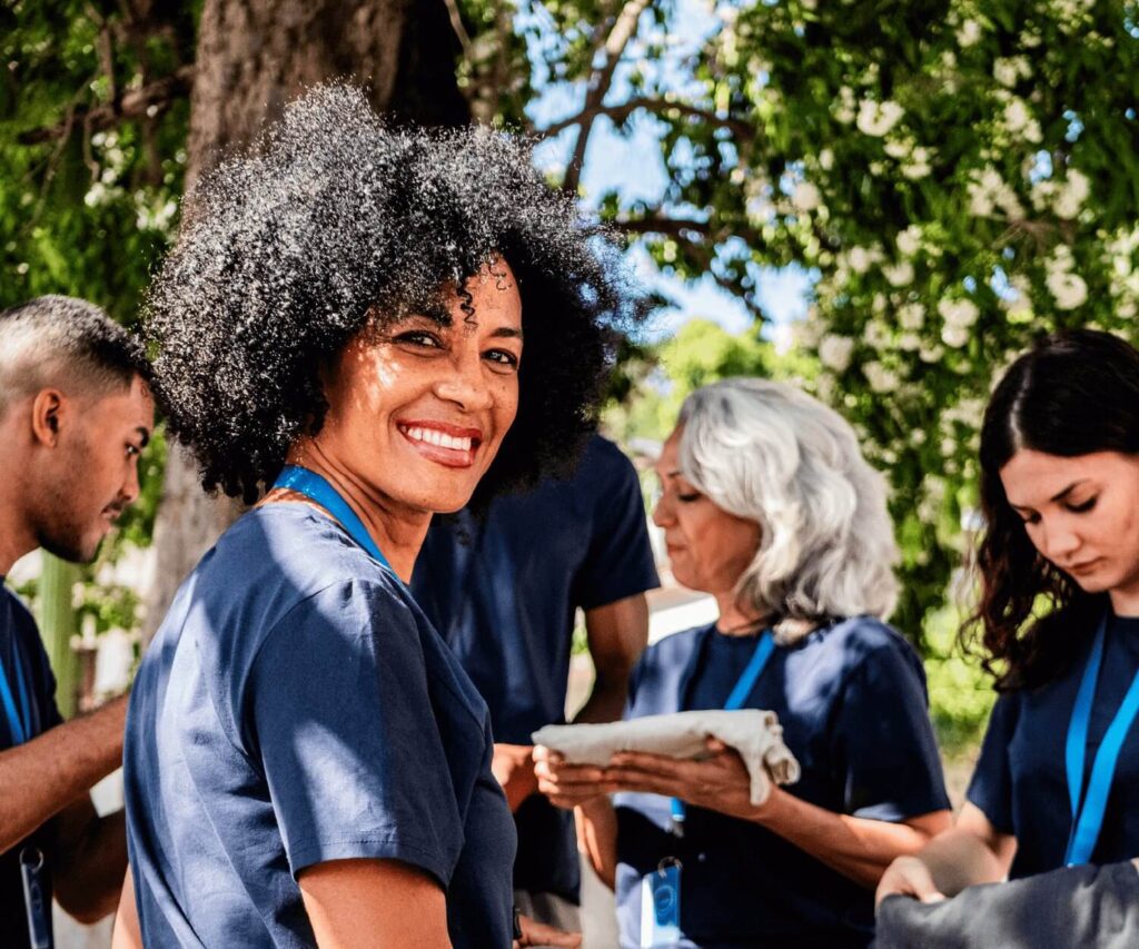 A group of volunteers wearing matching navy blue shirts gather outdoors. A smiling woman with curly hair in the foreground looks toward the camera, while other volunteers beside her focus on folding and preparing items. Trees and greenery provide a bright natural background.
