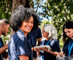 A group of volunteers wearing matching navy blue shirts gather outdoors. A smiling woman with curly hair in the foreground looks toward the camera, while other volunteers beside her focus on folding and preparing items. Trees and greenery provide a bright natural background.