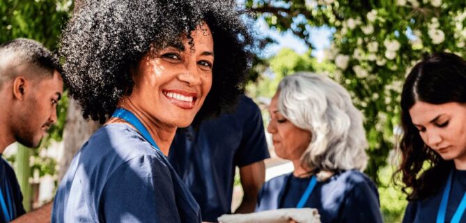 A group of volunteers wearing matching navy blue shirts gather outdoors. A smiling woman with curly hair in the foreground looks toward the camera, while other volunteers beside her focus on folding and preparing items. Trees and greenery provide a bright natural background.