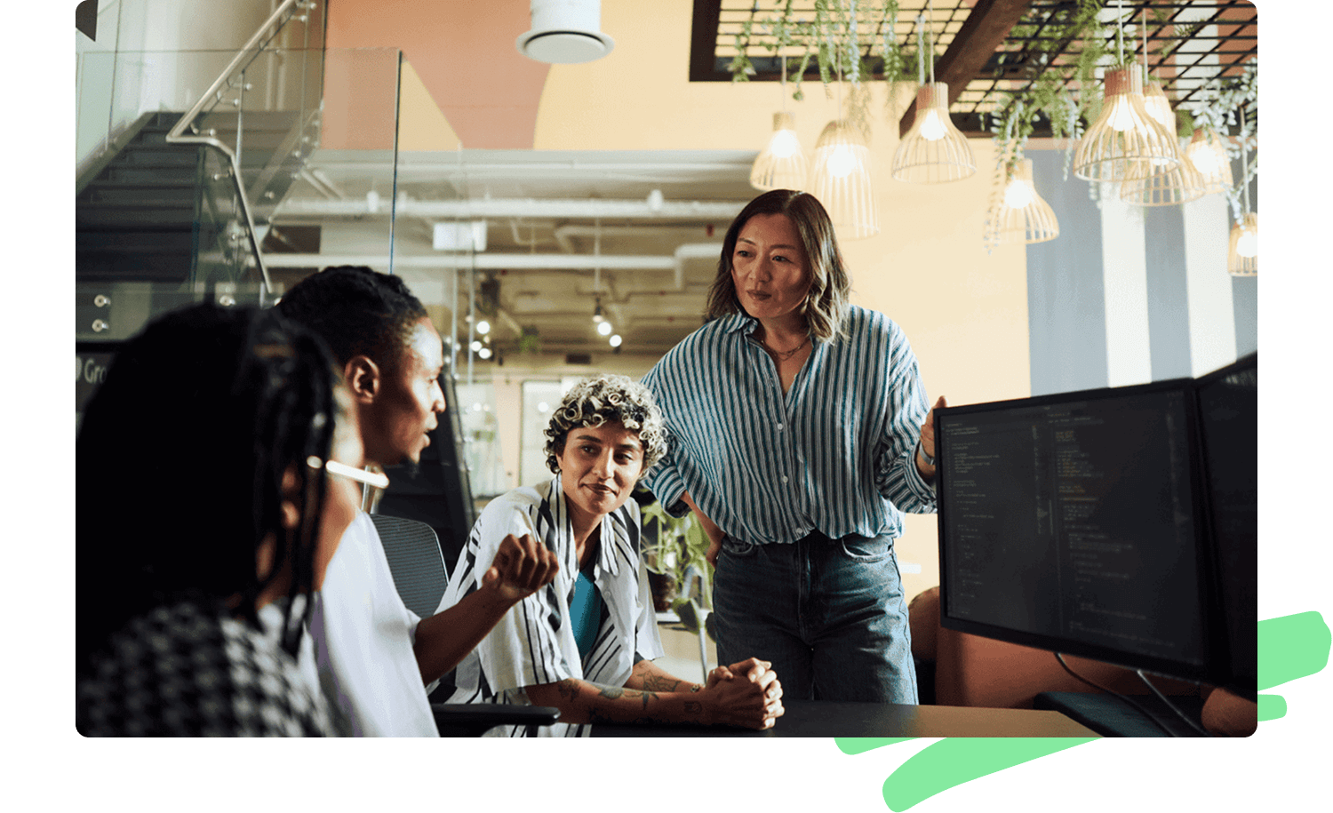 A diverse group of colleagues gathers around a desk with computer monitors displaying code. One woman stands, speaking to the team, while others listen attentively, symbolizing ethical discussions in technology development.