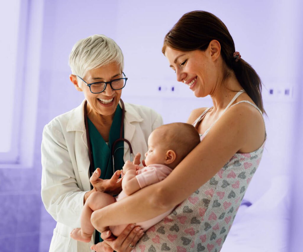 A smiling doctor with short gray hair and glasses stands beside a mother holding her baby. The doctor engages warmly with the baby while the mother looks on with a smile. The background has a soft purple overlay.