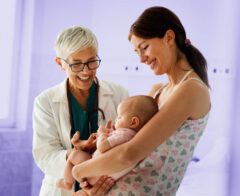 A smiling doctor with short gray hair and glasses stands beside a mother holding her baby. The doctor engages warmly with the baby while the mother looks on with a smile. The background has a soft purple overlay.