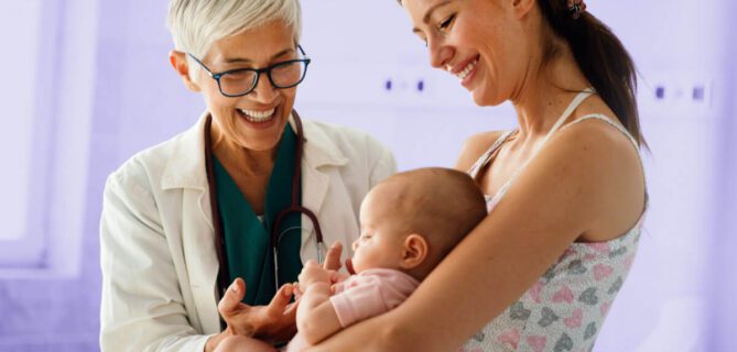 A smiling doctor with short gray hair and glasses stands beside a mother holding her baby. The doctor engages warmly with the baby while the mother looks on with a smile. The background has a soft purple overlay.