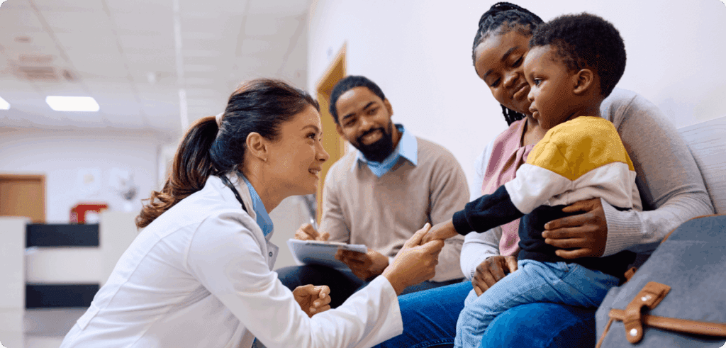 A doctor kneels down to greet a young boy sitting on his mother’s lap in a clinic waiting area. A man with a notepad sits beside them, smiling.