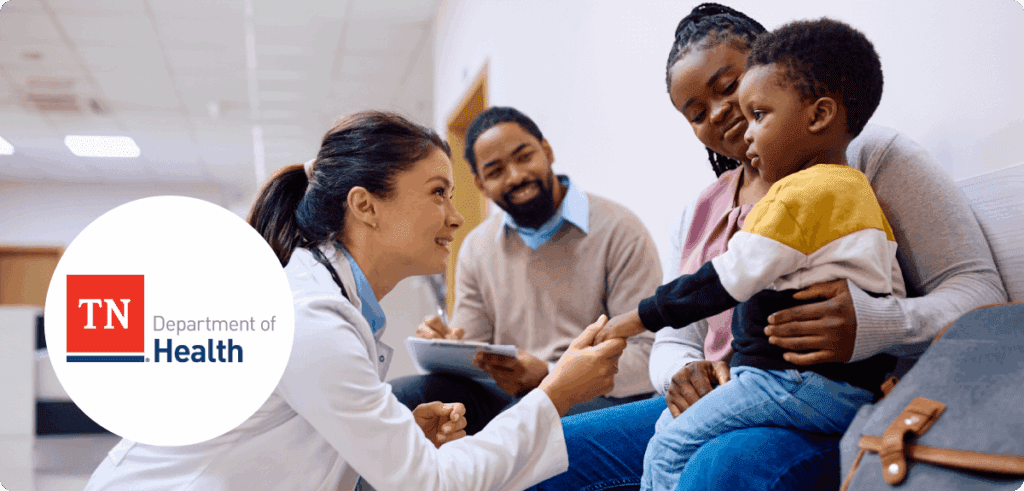 A doctor kneels down to greet a young boy sitting on his mother’s lap in a clinic waiting area. A man with a notepad sits beside them, smiling. On the left is the Tennessee Department of Health logo in a white circle.
