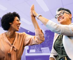 Two smiling colleagues sit indoors, leaning toward each other with joy as they exchange a high-five. One wears a peach blouse, the other a light blazer with glasses. The background shows an office space with a purple overlay.
