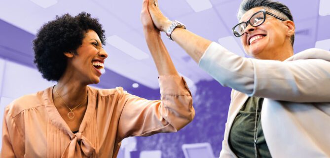 Two smiling colleagues sit indoors, leaning toward each other with joy as they exchange a high-five. One wears a peach blouse, the other a light blazer with glasses. The background shows an office space with a purple overlay.