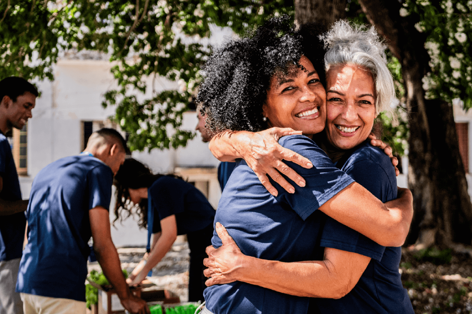 Two women wearing matching navy volunteer shirts hug each other and smile warmly while standing outdoors near other volunteers who are preparing supplies at tables. Trees and sunlight frame the background, giving a sense of community and teamwork.