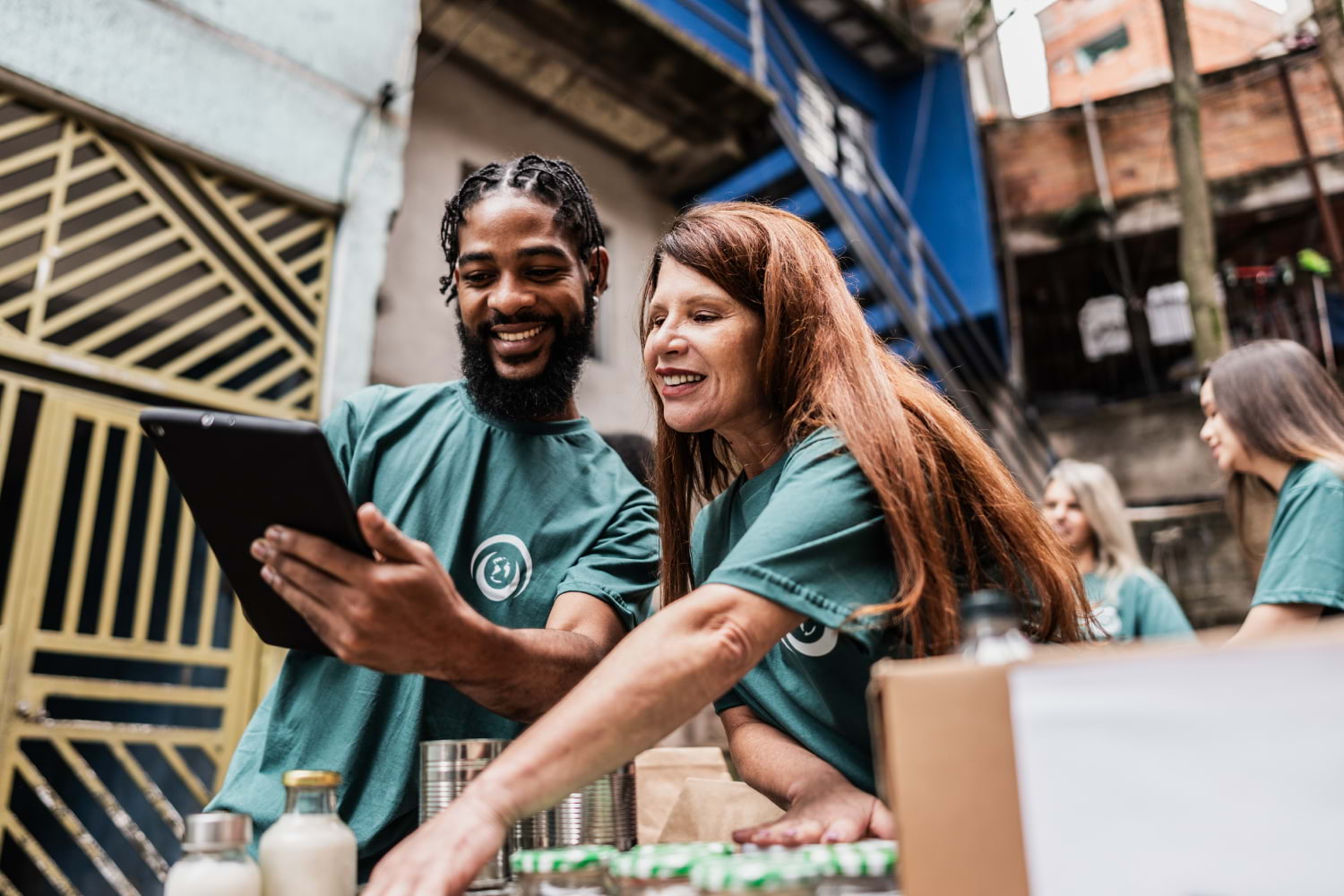 A man and woman wearing green volunteer shirts work together outdoors at a donation or food distribution event. The man holds a tablet while smiling, and the woman reaches toward a table with canned goods and supplies. Other volunteers are visible in the background.