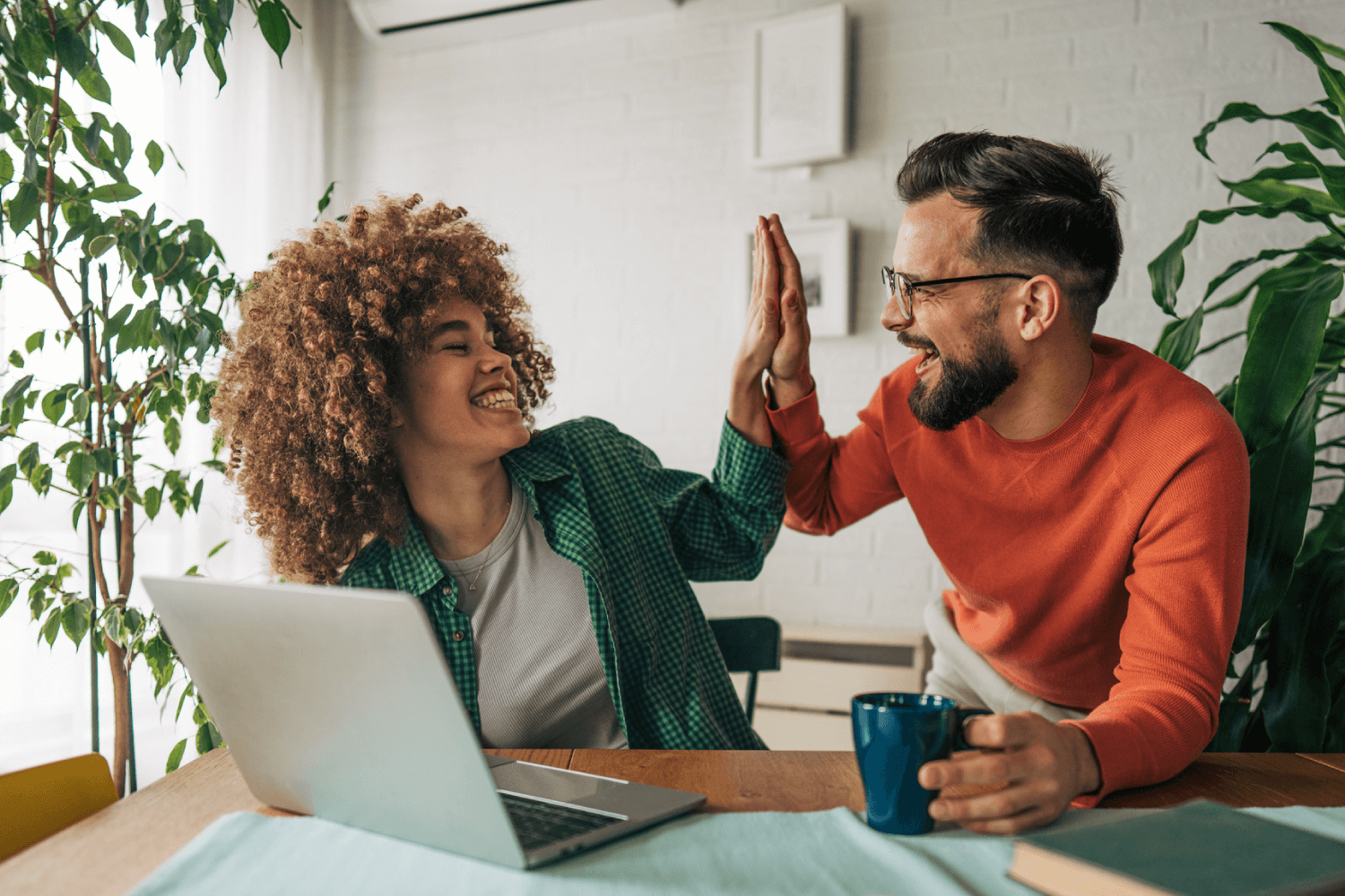 Two colleagues sit together at a table with a laptop and coffee mug, smiling as they high-five each other. The background shows bright natural light and houseplants, creating a cheerful and collaborative atmosphere.