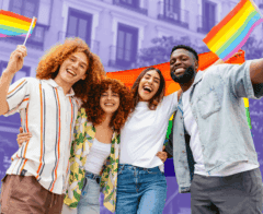 A diverse group of four friends stand together outdoors, smiling and holding rainbow Pride flags. They appear joyful and united, with a rainbow flag stretched behind them and buildings in the background.