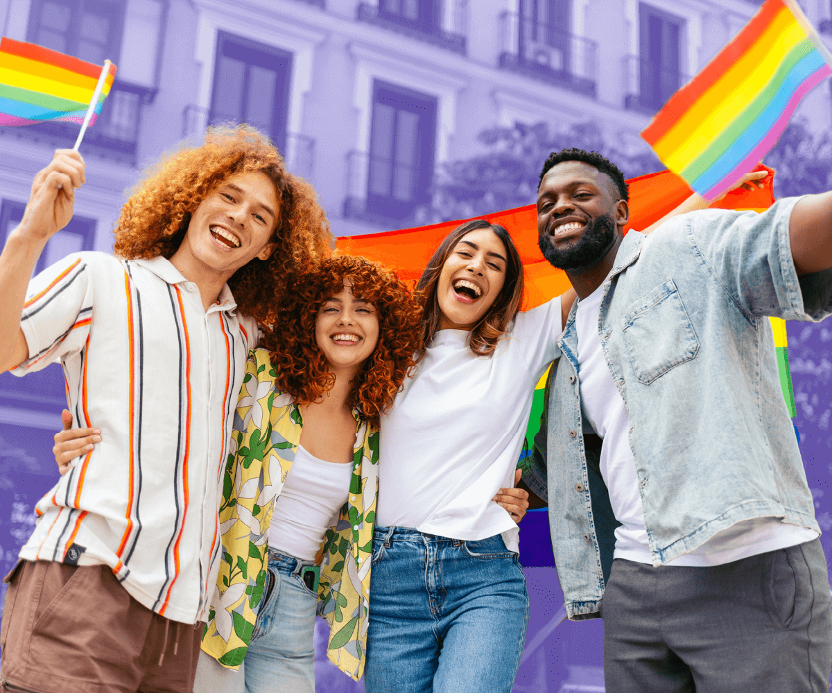 A diverse group of four friends stand together outdoors, smiling and holding rainbow Pride flags. They appear joyful and united, with a rainbow flag stretched behind them and buildings in the background.
