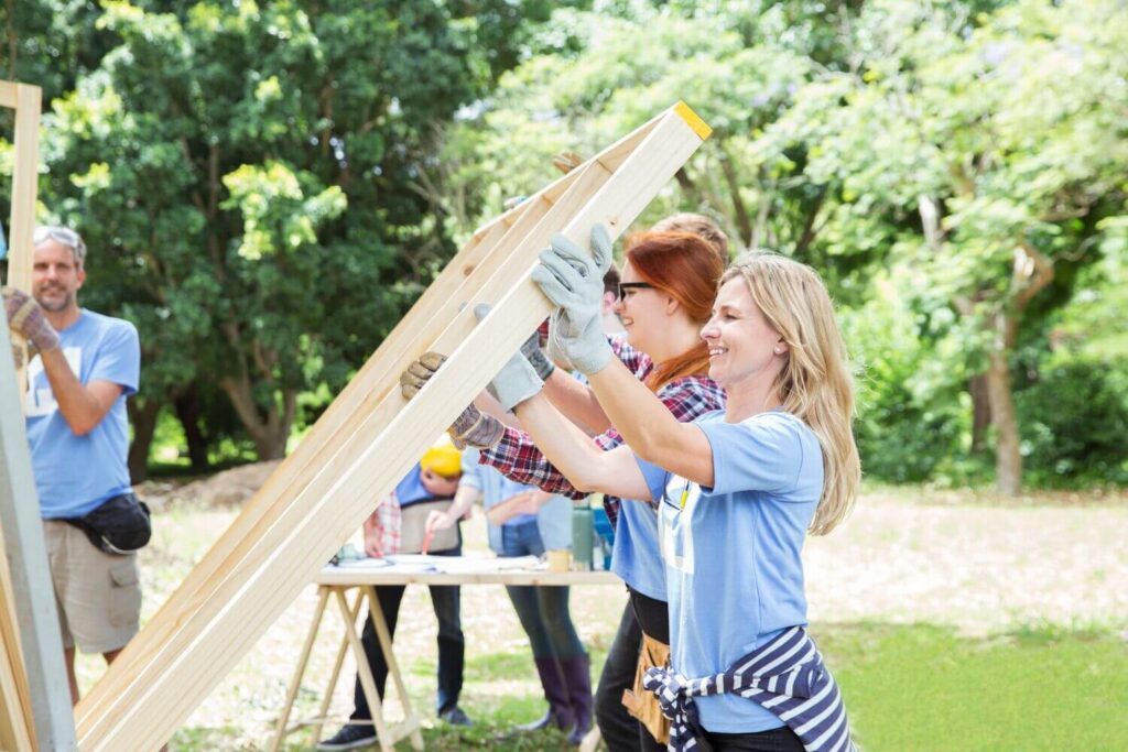 A group of volunteers outdoors work together to raise a wooden frame, likely as part of a construction or community building project. Smiling participants wear gloves and casual clothing, with trees and other volunteers visible in the background.