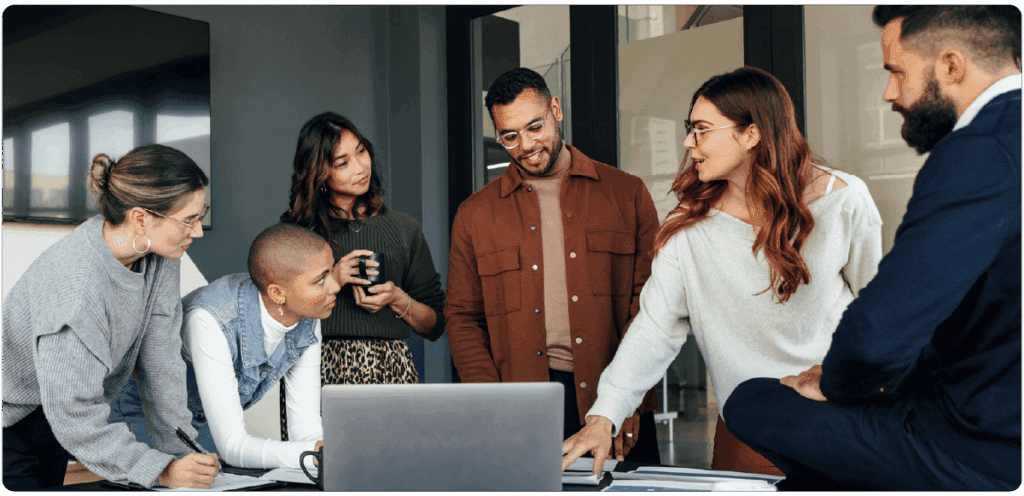A group of five professionals gathers around a table with a laptop, reviewing a project together. One woman points at the screen while others listen attentively and smile, symbolizing teamwork and collaboration in a modern office environment.