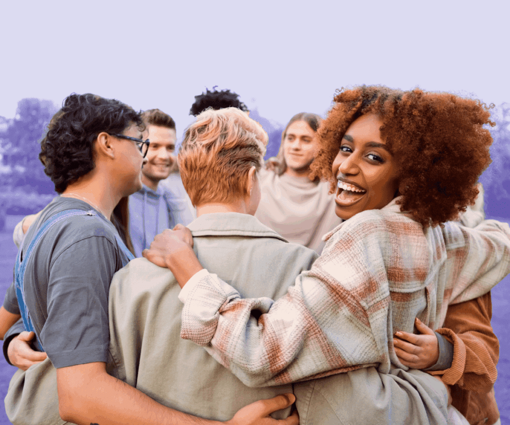 A diverse group of people stands outdoors in a circle with arms around each other, smiling and laughing together. One woman turns toward the camera with a joyful expression. The background has a soft purple tint, evoking community and connection.