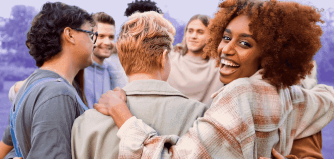 A diverse group of people stands outdoors in a circle with arms around each other, smiling and laughing together. One woman turns toward the camera with a joyful expression. The background has a soft purple tint, evoking community and connection.