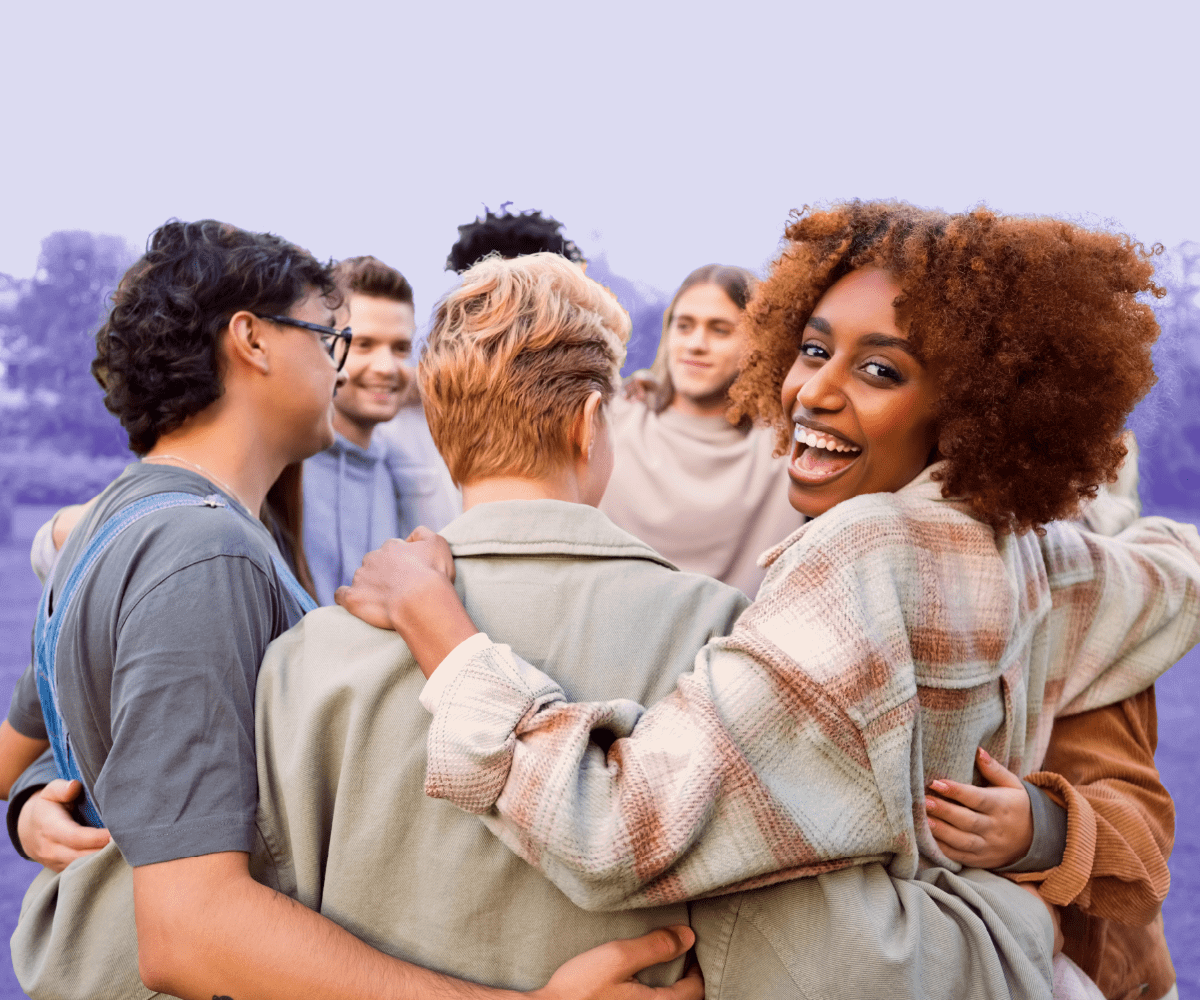 A diverse group of people stands outdoors in a circle with arms around each other, smiling and laughing together. One woman turns toward the camera with a joyful expression. The background has a soft purple tint, evoking community and connection.