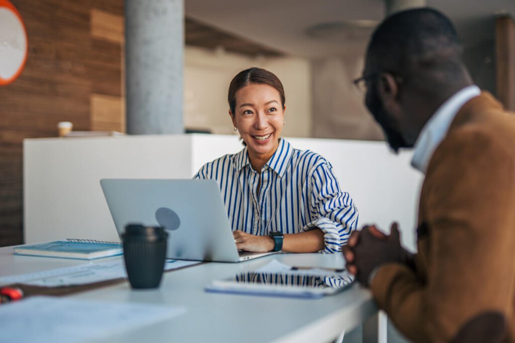 A woman smiling and talking with a colleague across a table in a modern office. She has a laptop open in front of her, and they appear to be having a friendly, productive conversation.