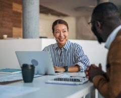 A woman smiling and talking with a colleague across a table in a modern office. She has a laptop open in front of her, and they appear to be having a friendly, productive conversation.
