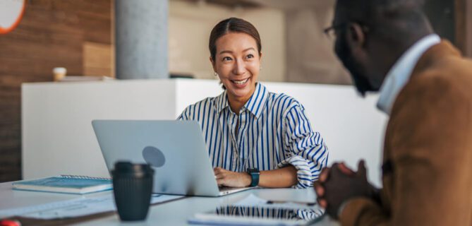A woman smiling and talking with a colleague across a table in a modern office. She has a laptop open in front of her, and they appear to be having a friendly, productive conversation.